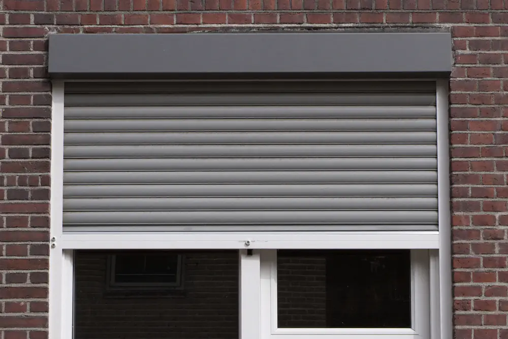 Wit kozijn met een grijs rolluik en rolluikkast aan de buitenkant van een baksteenwoning